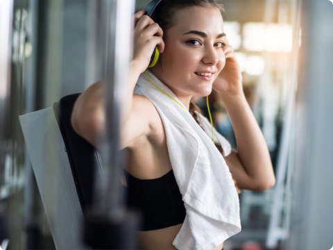 一位身穿黑色运动内衣的年轻女子坐在健身房的健身器材上，微笑着调整耳机。她的脖子上搭着一条白色毛巾，似乎正在休息。背景是健身器材，阳光透过窗户照射进来。