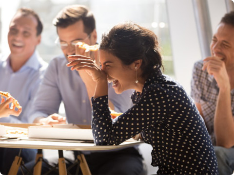 一群同事在明亮的办公室里吃着披萨享受着轻松愉快的时刻。前景中一位身着波点衬衫的女性拿着一块披萨开怀大笑。其他同事身着商务休闲装，面带微笑，围桌交谈。