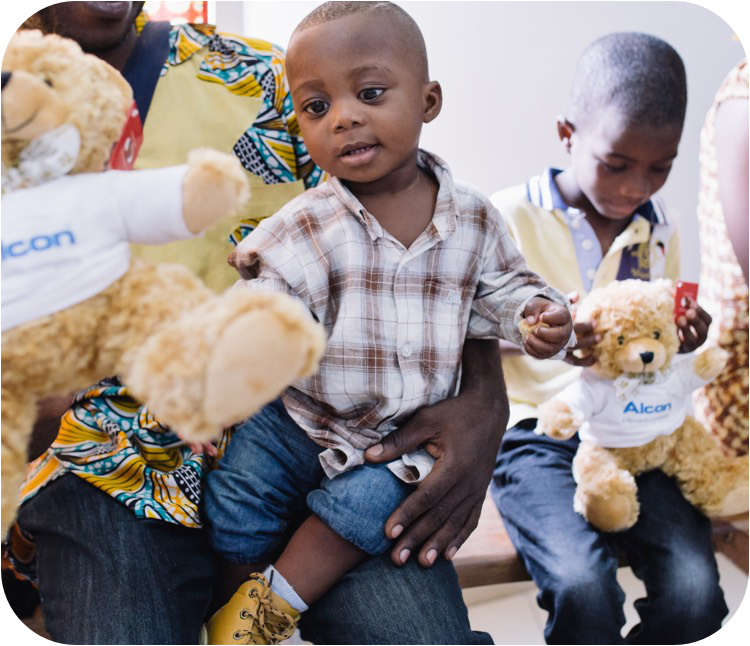 一个小孩坐在成年人的腿上，穿着格子衬衫和卷起的牛仔裤，正伸手去拿一只毛绒泰迪熊，泰迪熊的衬衫上印有 Alcon 标识。背景中的另一个孩子也抱着一只类似的泰迪熊，坐在那里看着小熊。