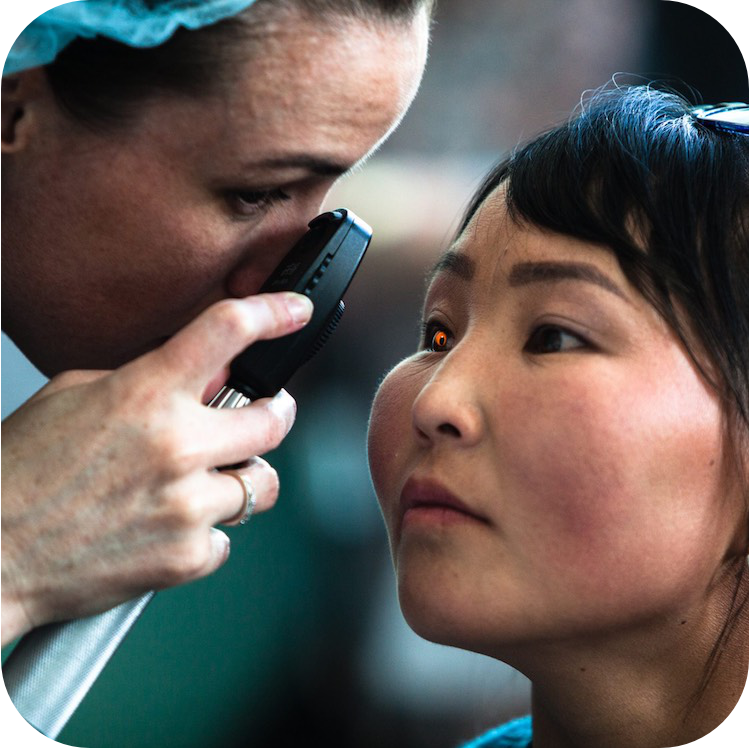 眼科专业人员使用手持式眼底镜检查患者的眼睛。女患者直视前方，专业人员则俯身进行详细的眼部检查。眼底镜发出微弱的橙色光，在病人的眼睛里反射出来。