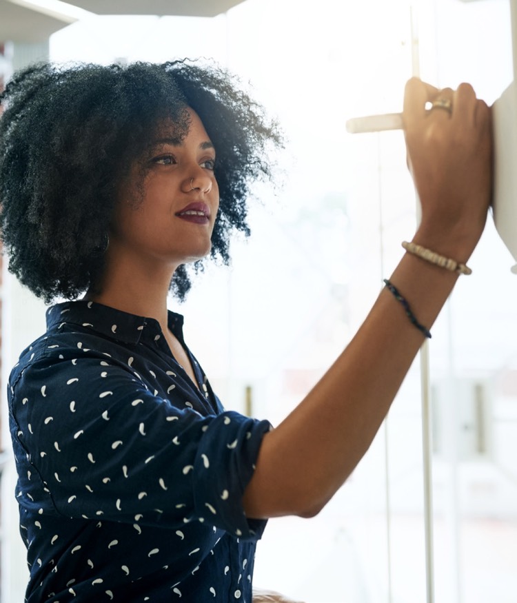 在一个光线充足的空间里，一位卷发、身着深蓝色带图案衬衫的女性在白板上写字或画画。