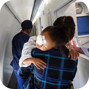 一名眼睛上绑着纱布的小孩正趴在一名妇女的肩头休息，妇女穿着格子衬衫，抱着他穿过一家医疗机构的狭窄走廊，走廊上还能看到一名穿手术服的医疗工作者。这场景表明小孩刚刚接受了眼部手术，正处于术后护理或恢复阶段。